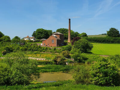 Terracotta brick building with tall brick chimney surrounded by green lush grass, trees and fields.