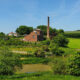 Terracotta brick building with tall brick chimney surrounded by green lush grass, trees and fields.