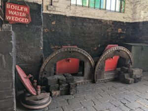 Black, grey and red metal work from front of Lancashire Boiler piled on grey stone floor.