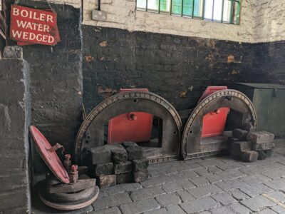 Black, grey and red metal work from front of Lancashire Boiler piled on grey stone floor.